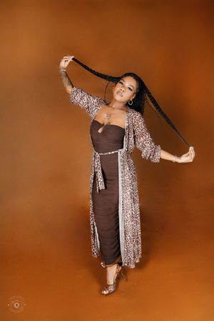 A woman in a brown midi dress and leopard print kimono poses confidently, holding her long braids up against a warm brown background.