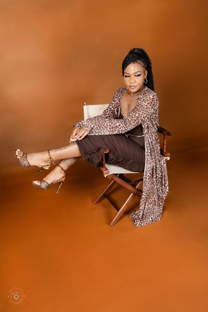 Woman in leopard print outfit and strappy heels seated on a director's chair against a warm brown background.