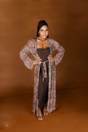 Woman in brown dress and leopard print duster poses confidently against warm brown background.