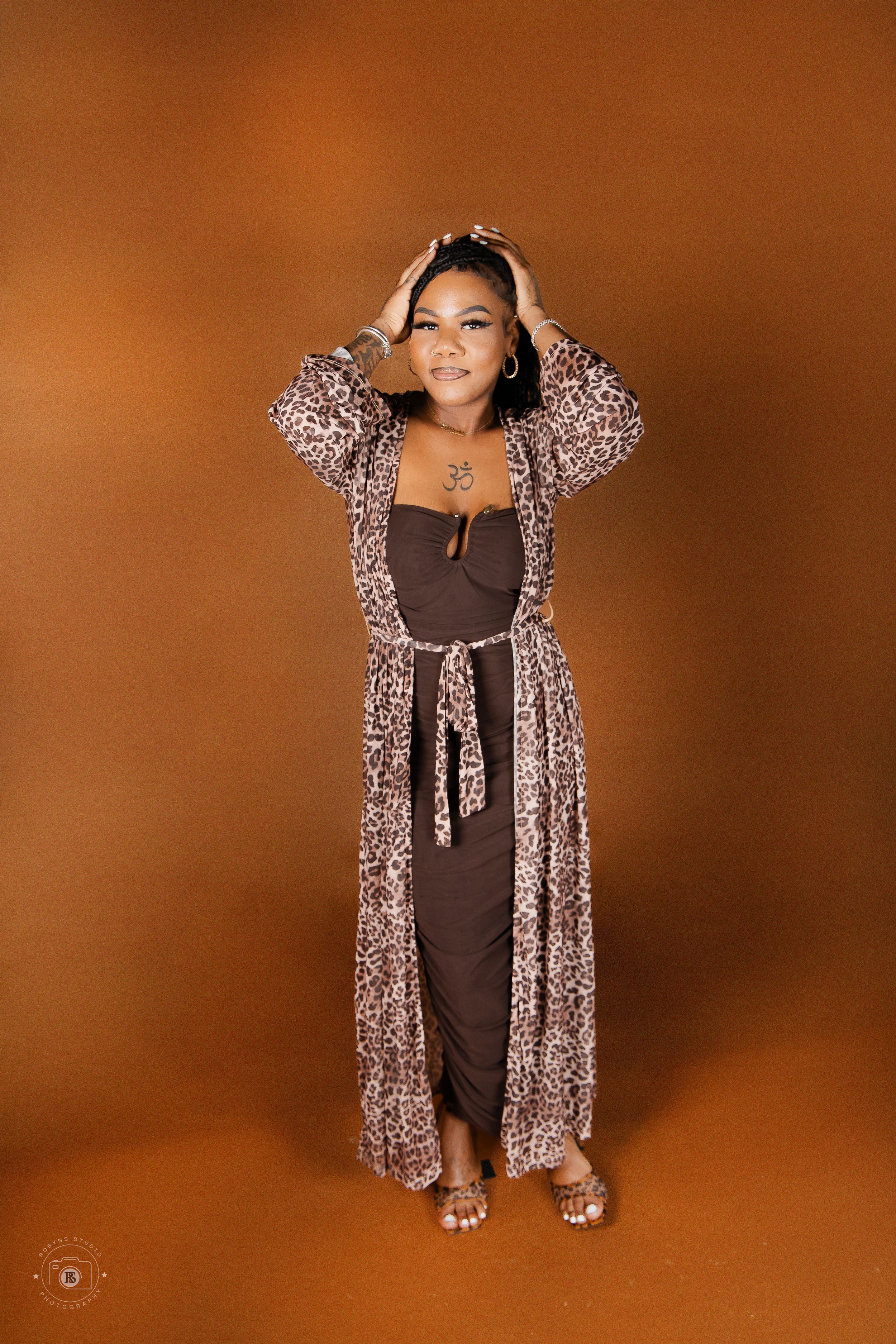 Woman in brown jumpsuit and leopard print duster poses confidently against warm orange backdrop.