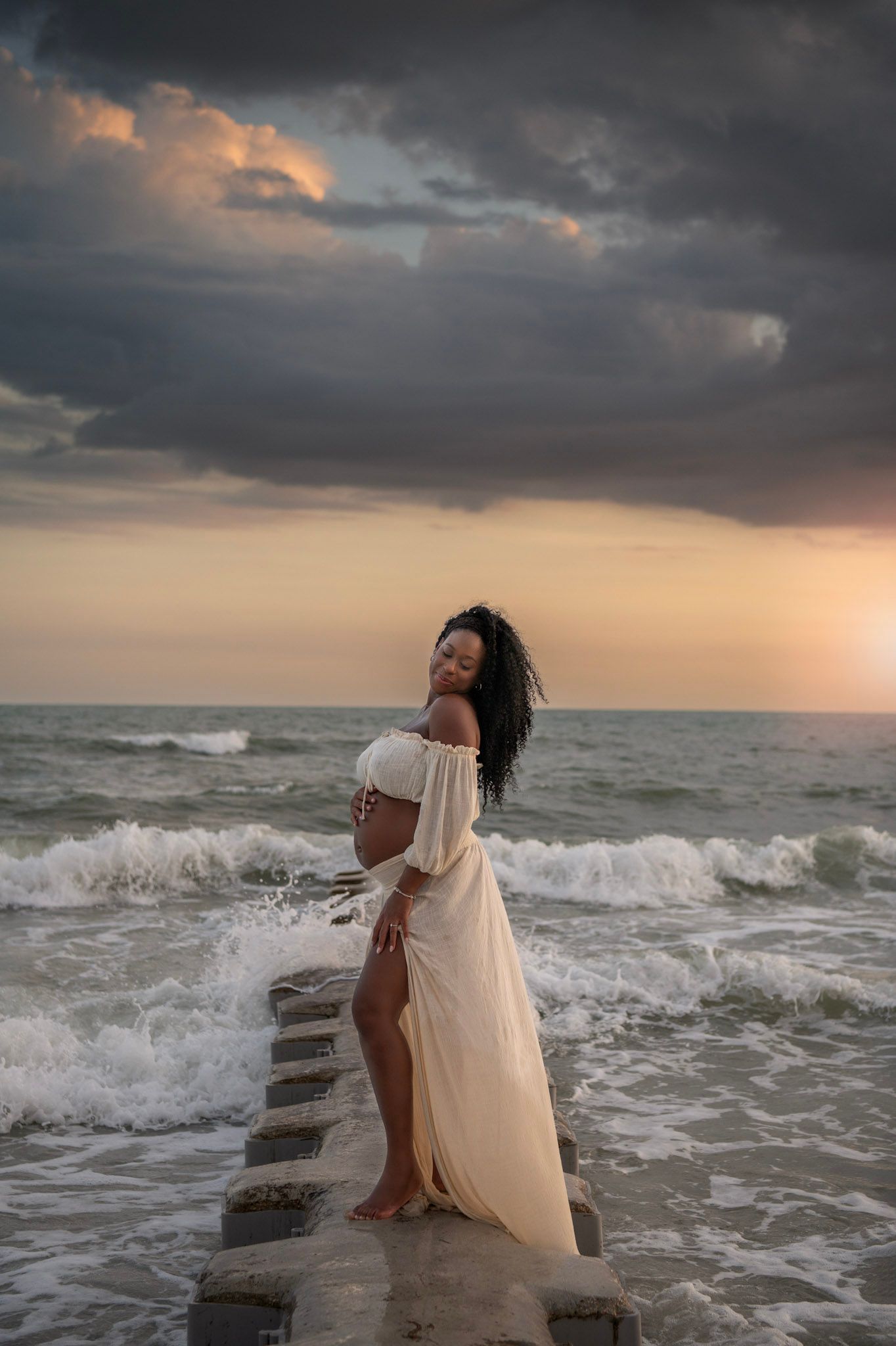 Pregnant woman in a flowing white dress standing on a pier amid ocean waves at sunset.
