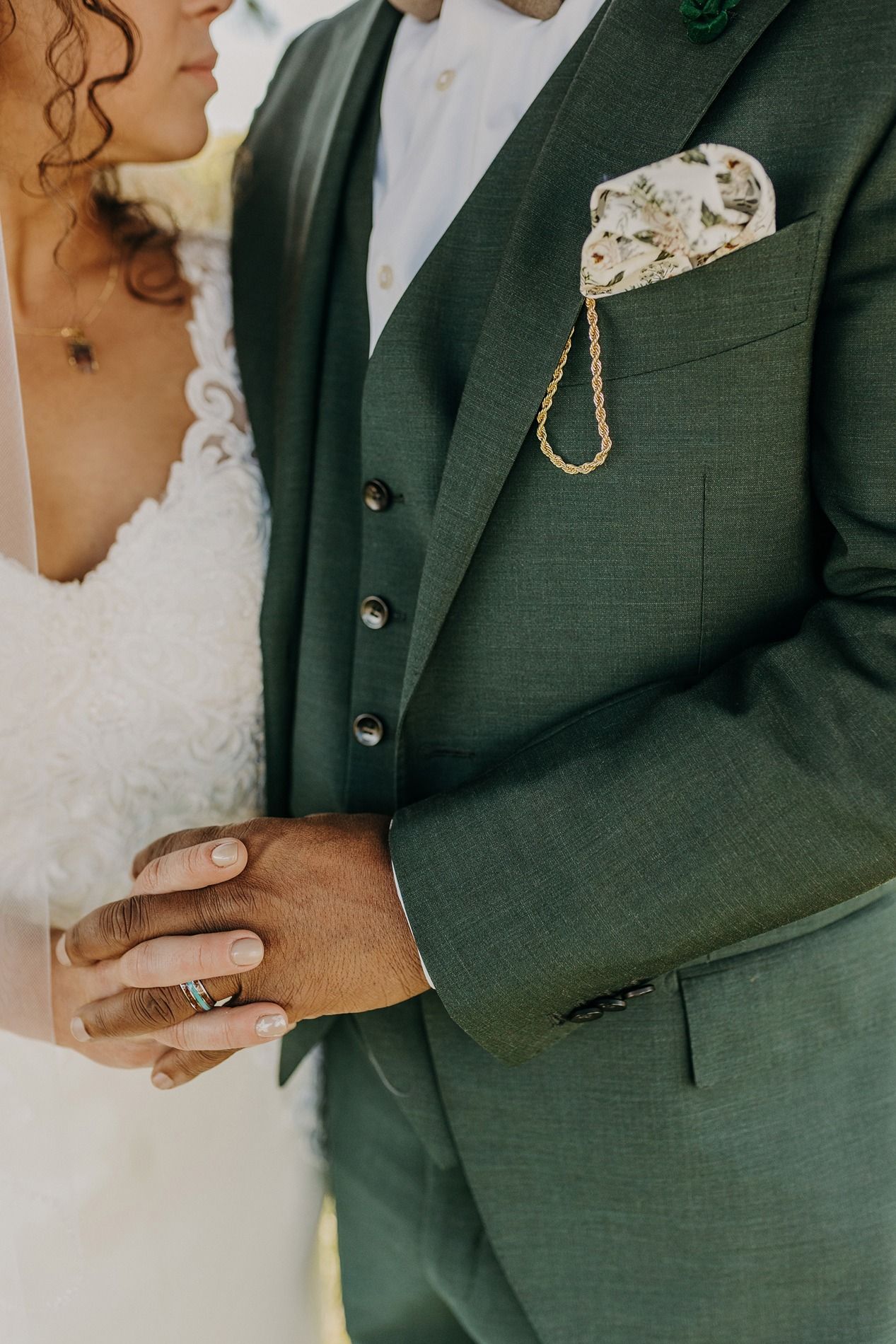Bride and groom in elegant green suit with floral pocket square and gold chain at wedding ceremony.