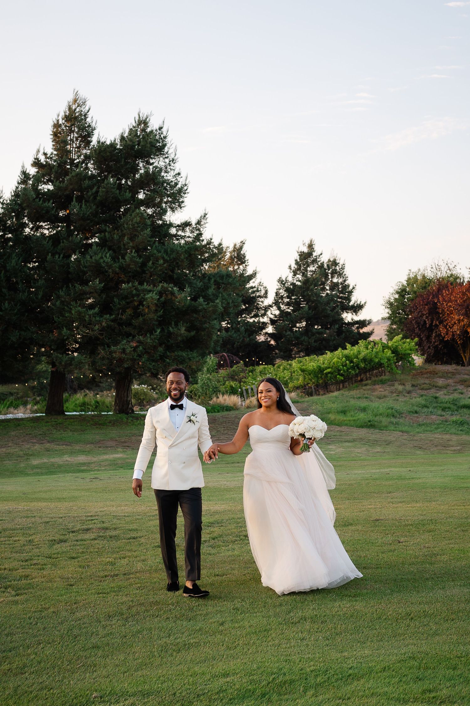 A joyful bride and groom walk hand-in-hand on a lush green lawn, she in a white gown, he in a white tuxedo jacket.