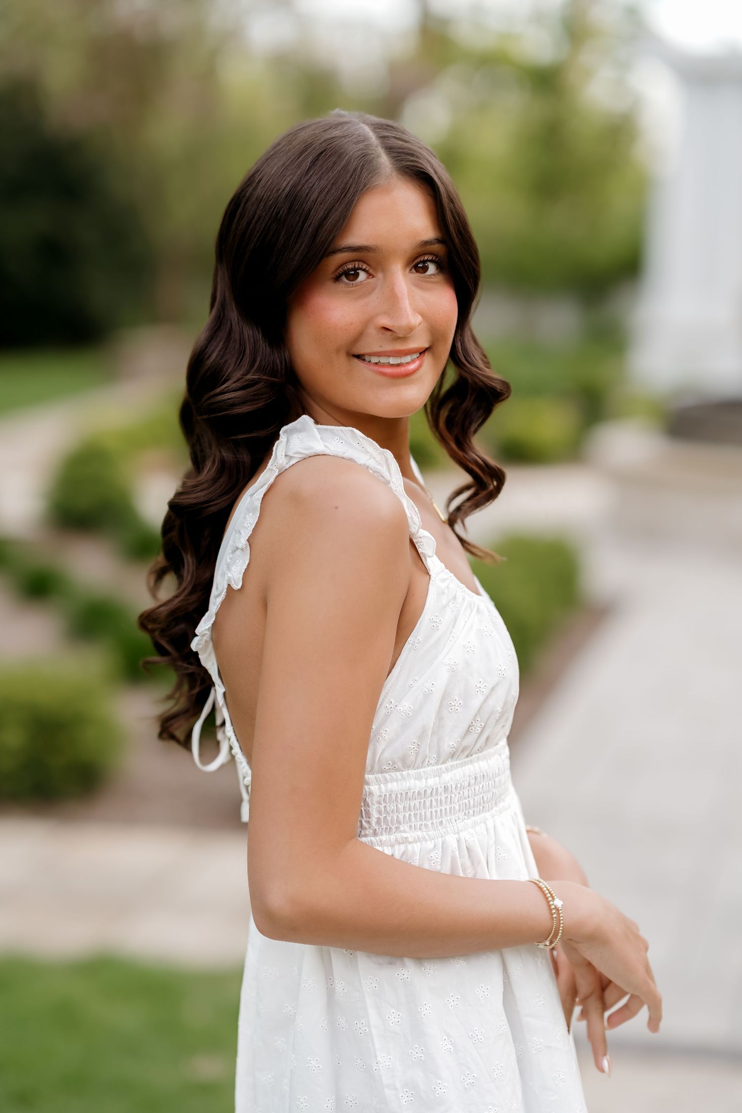 Young woman in white sundress with ruffled straps smiling outdoors in a garden setting.