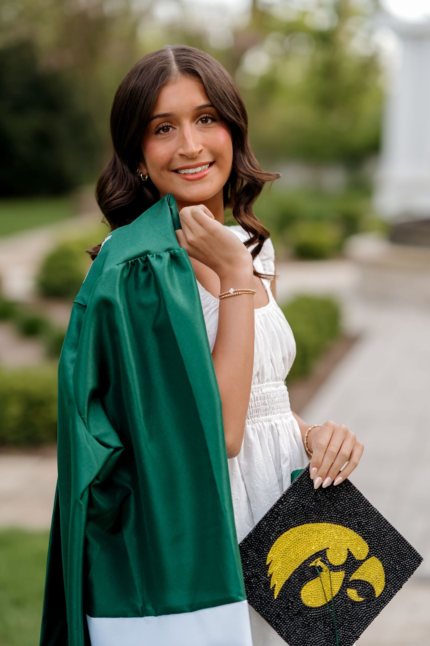 Smiling young woman in green graduation gown holding University of Iowa decorated cap outdoors.