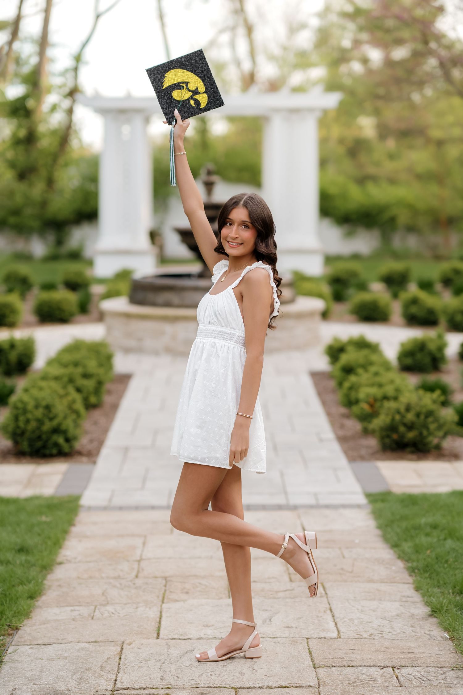 Graduate in white dress holding Iowa Hawkeyes cap aloft on garden pathway with columns.