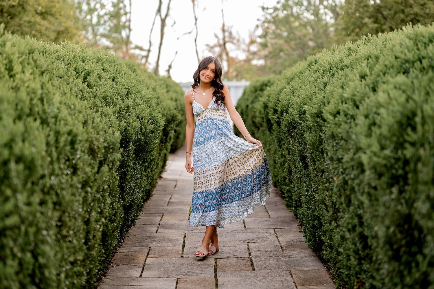 Woman in flowing blue patterned maxi dress walking through lush green hedge pathway.