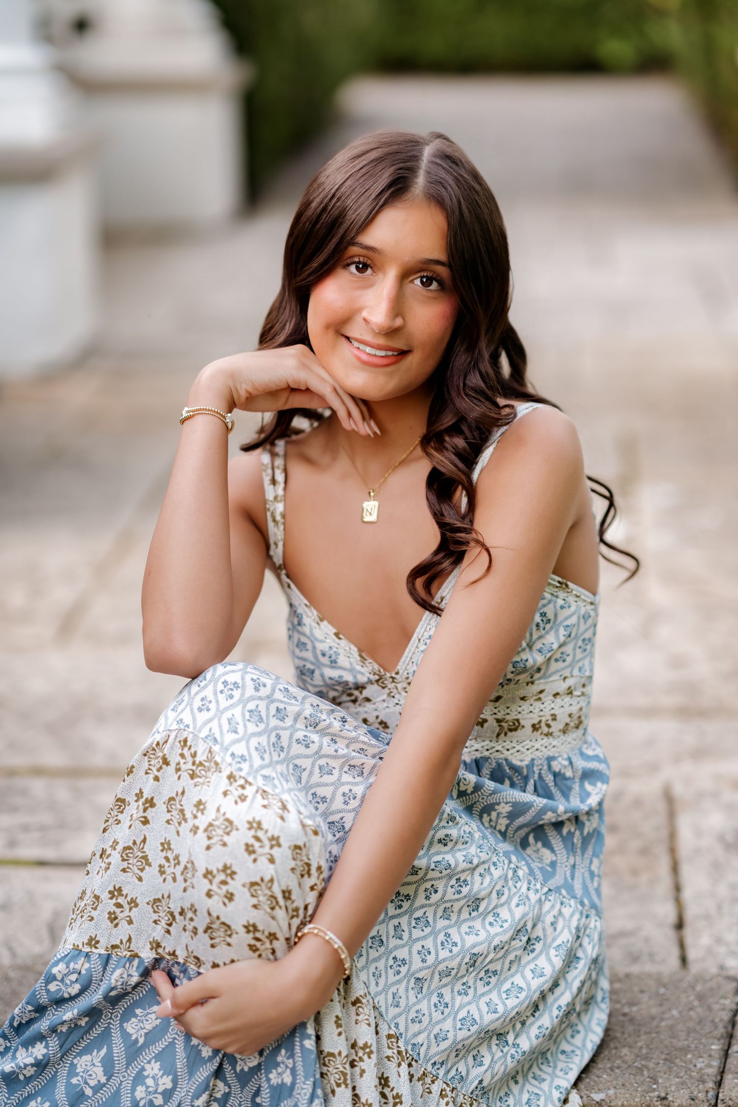 Young woman in blue floral sundress sitting on stone steps smiling elegantly outdoors.
