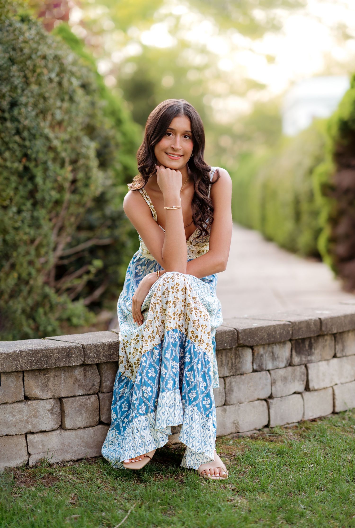 Young woman in floral maxi dress sitting on stone wall in lush garden pathway, smiling gracefully.