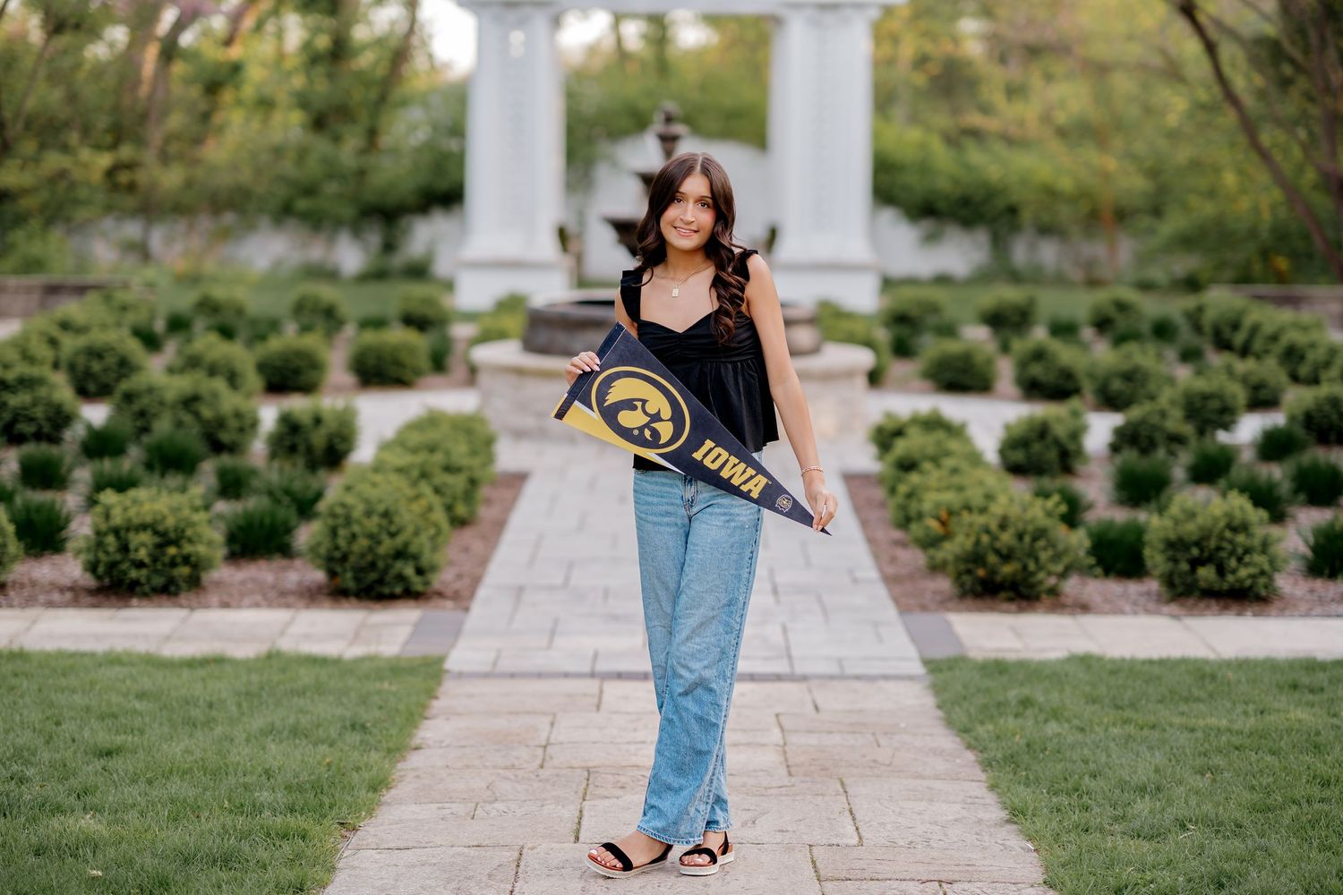 Young woman holding University of Iowa pennant poses on garden pathway in jeans and black top.