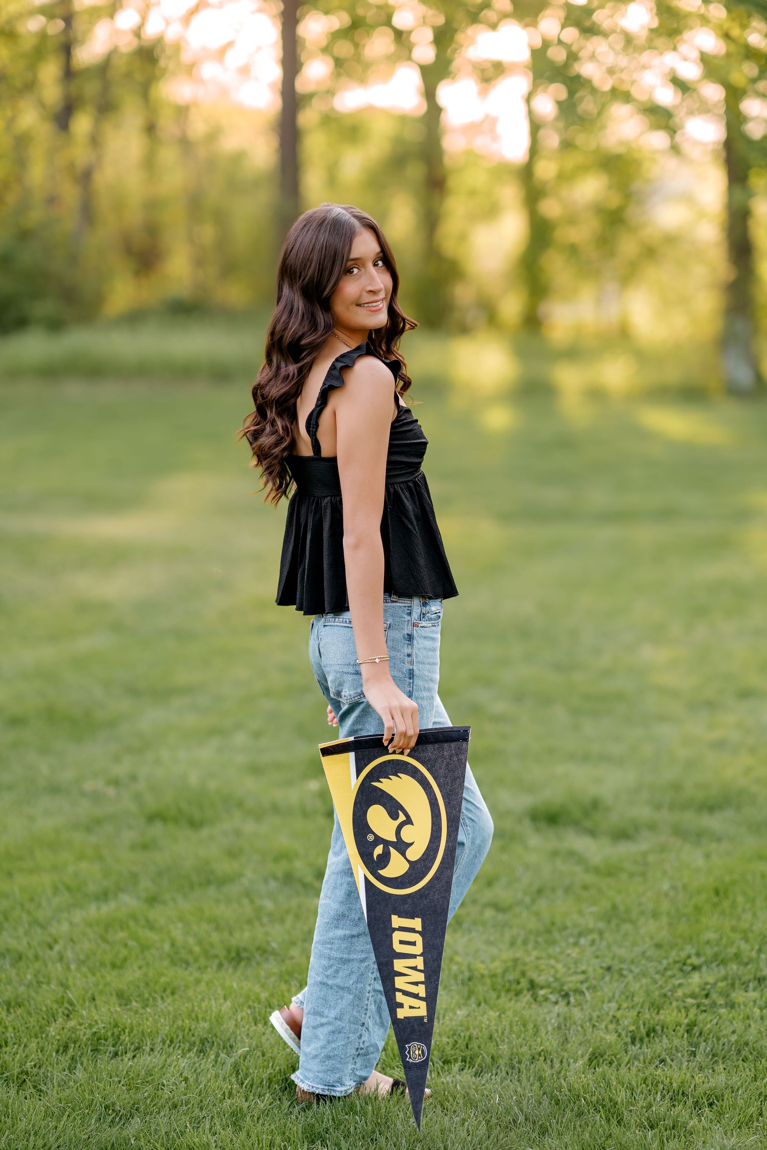 Young woman holding an Iowa Hawkeyes pennant flag, wearing black top and jeans, posing outdoors on green grass.