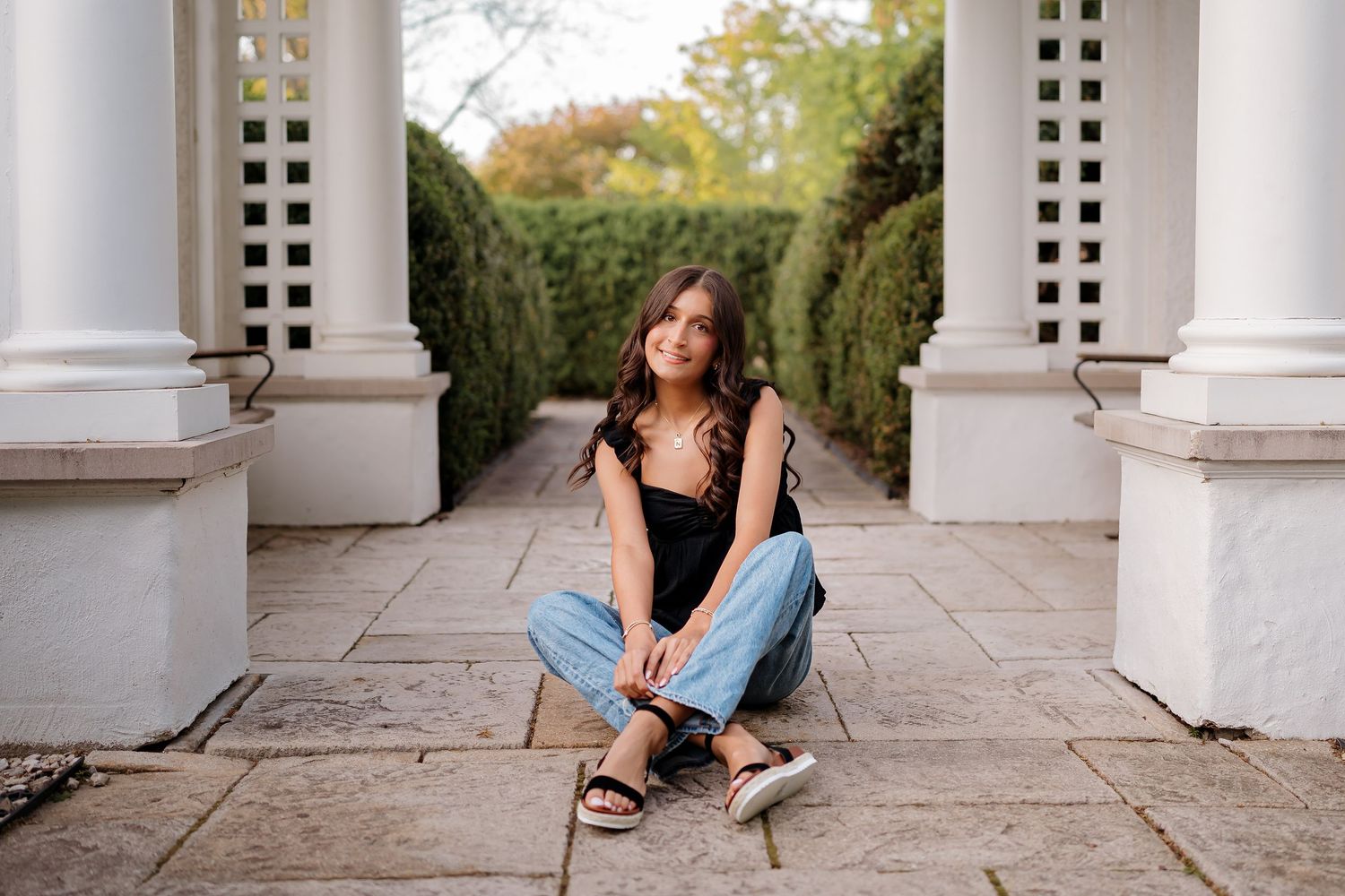Young woman in jeans and black top sitting on stone patio between white columns with green hedges backdrop.