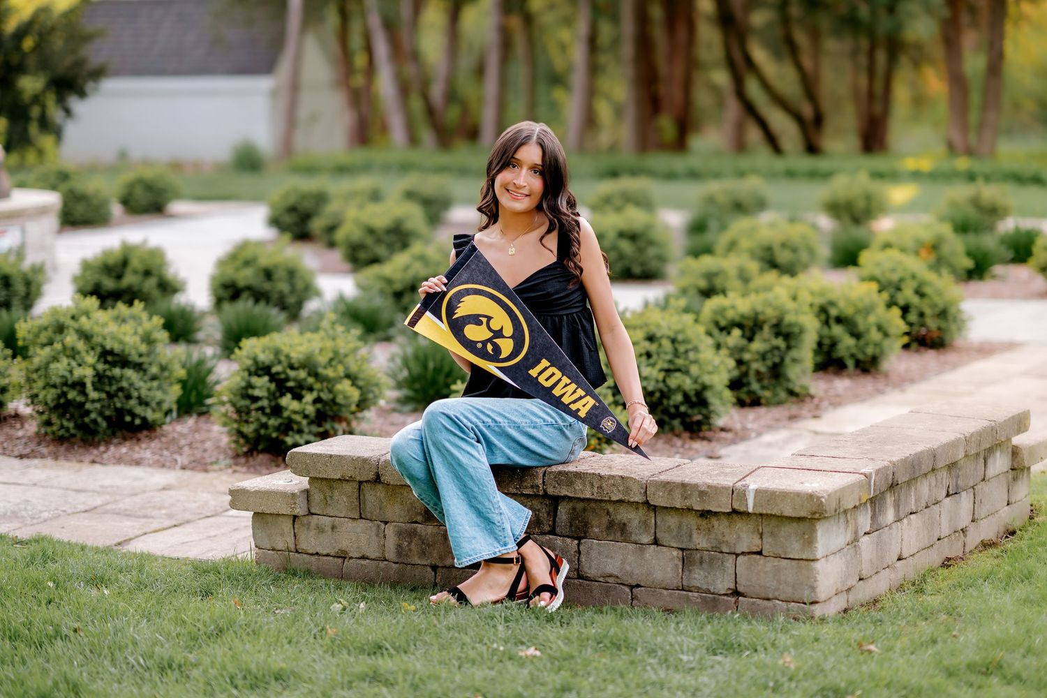 Young woman sitting on stone wall holding University of Iowa pennant flag in lush garden setting.
