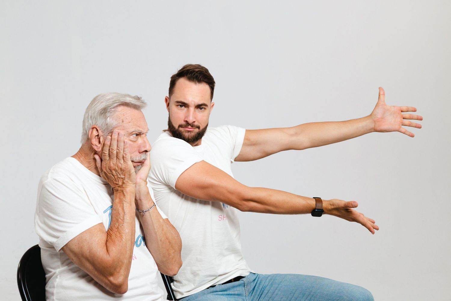 Young man in white t-shirt gesturing with outstretched arm beside shocked older man holding his face.