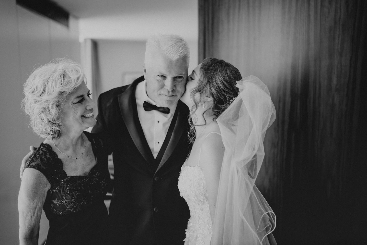 Black and white wedding photo of bride in veil kissing groom's cheek while older woman smiles beside them.