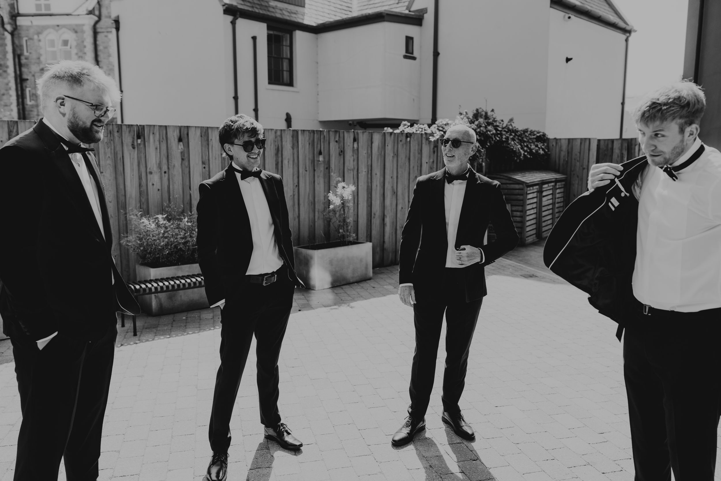 Four groomsmen in black tuxedos and bow ties gathered outdoors in black and white photo.