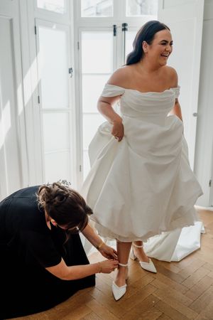 Bride in off-shoulder white wedding dress getting shoes fastened by bridesmaid.