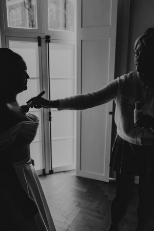 Black and white photo of bride being helped into her wedding dress by a bridesmaid near a window.