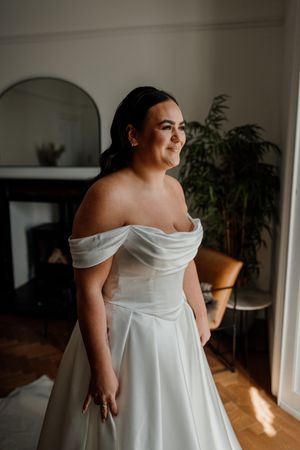 A bride in an elegant off-shoulder white satin gown poses in a sunlit room before her wedding.