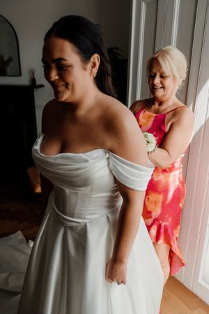 Woman in pink floral dress helping bride into elegant off-shoulder white wedding gown.