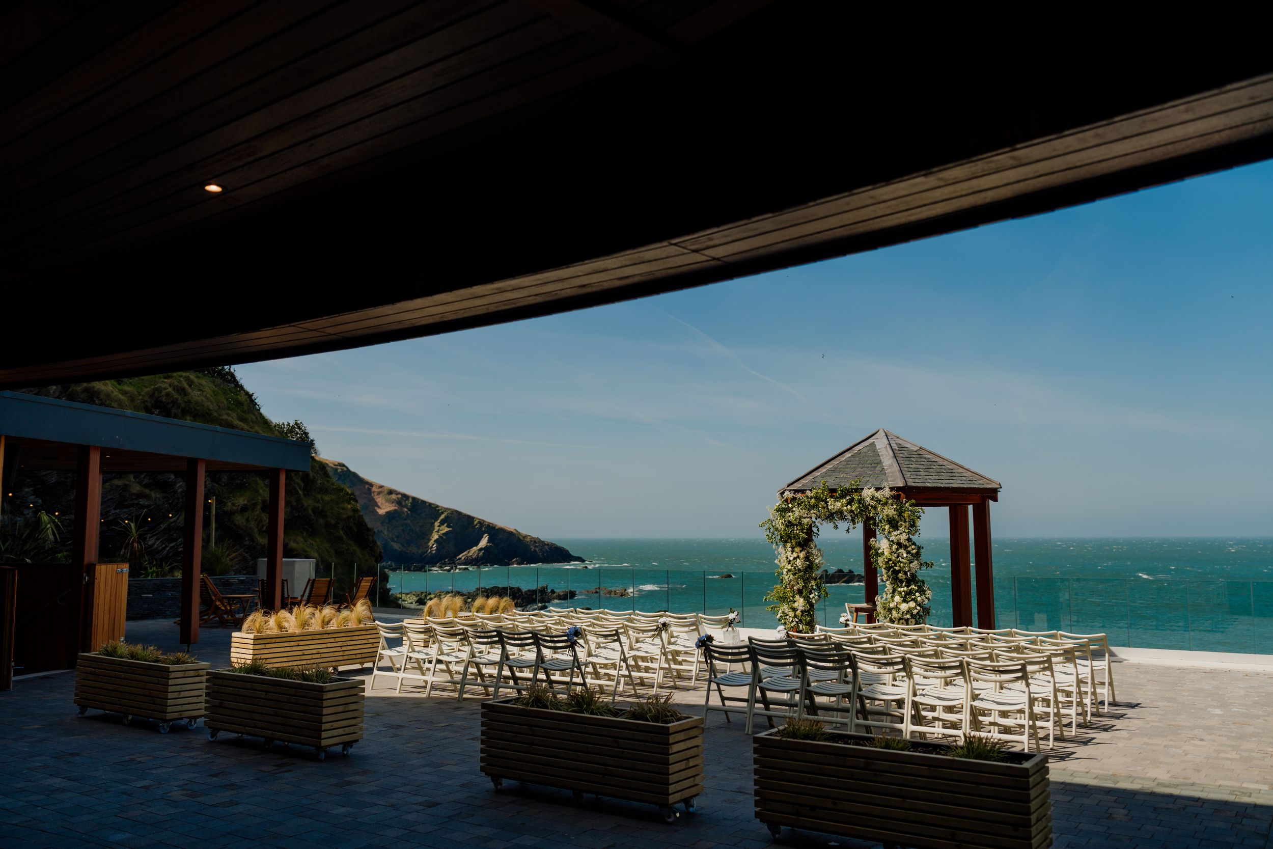 Elegant seaside wedding venue setup with white chairs, floral arch gazebo and ocean views.