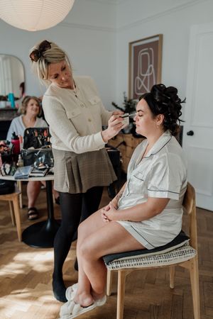 Makeup artist applying eyeshadow to a seated bride in pajamas during wedding preparations in a bright room.