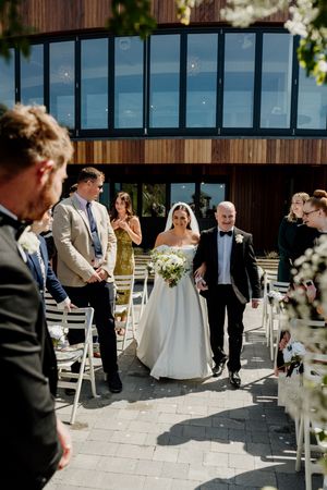 A bride walks down the outdoor aisle with her escort at a sunny modern wedding ceremony.