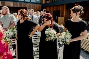Bridesmaids in black dresses holding white bouquets emotional during wedding ceremony aisle procession.