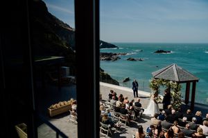 Aerial view of coastal wedding ceremony with couple at floral gazebo and seated guests.