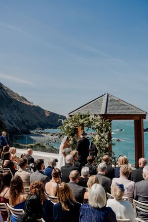 Outdoor coastal wedding ceremony with guests seated facing couple at floral arch overlooking ocean.