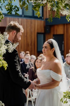 Bride and groom exchange vows outdoors, surrounded by white floral arrangements and wedding guests.