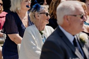 Elderly female wedding guest in white jacket and blue fascinator smiles during outdoor ceremony.