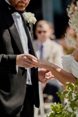 Groom placing wedding ring on brides finger during an outdoor wedding ceremony in bright sunlight.