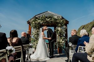 Bride and groom share a kiss at an outdoor coastal wedding ceremony beneath a floral arch gazebo.