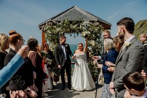 A joyful bride and groom celebrate at an outdoor coastal wedding ceremony with floral arch.