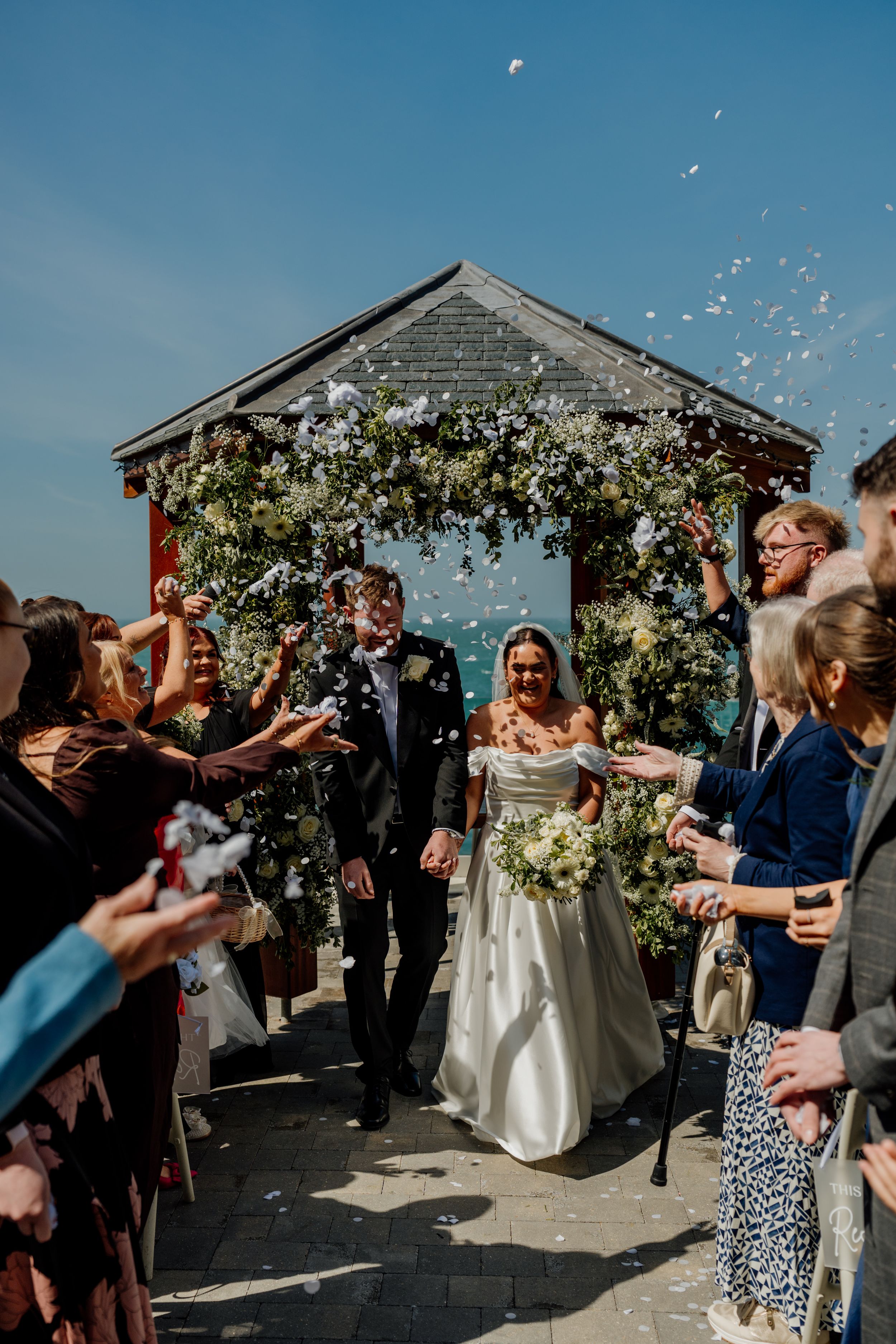 Bride and groom walk through confetti shower under floral arch with ocean backdrop on sunny wedding day.