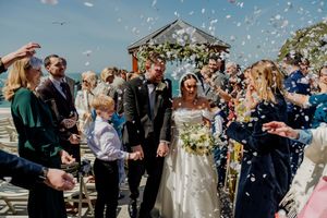 Bride and groom walk through colorful confetti shower surrounded by cheering wedding guests outdoors.
