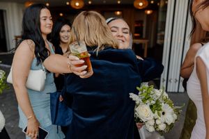 Joyful wedding guest hugging bride holding white bouquet at outdoor reception.