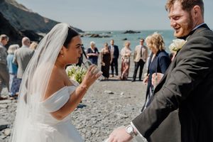 Bride laughing with bouquet while groom smiles on a rocky beach during their coastal wedding.