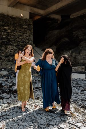 Three women in colorful dresses laughing on rocky beach at wedding celebration.