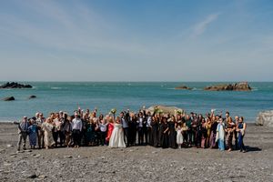 Large wedding party group photo on rocky beach with turquoise ocean backdrop.