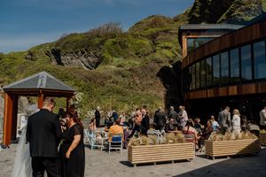Wedding guests mingle outdoors at a cliffside venue with ocean views on a sunny day.