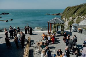 Aerial view of wedding guests mingling on coastal terrace with ocean and cliffs.