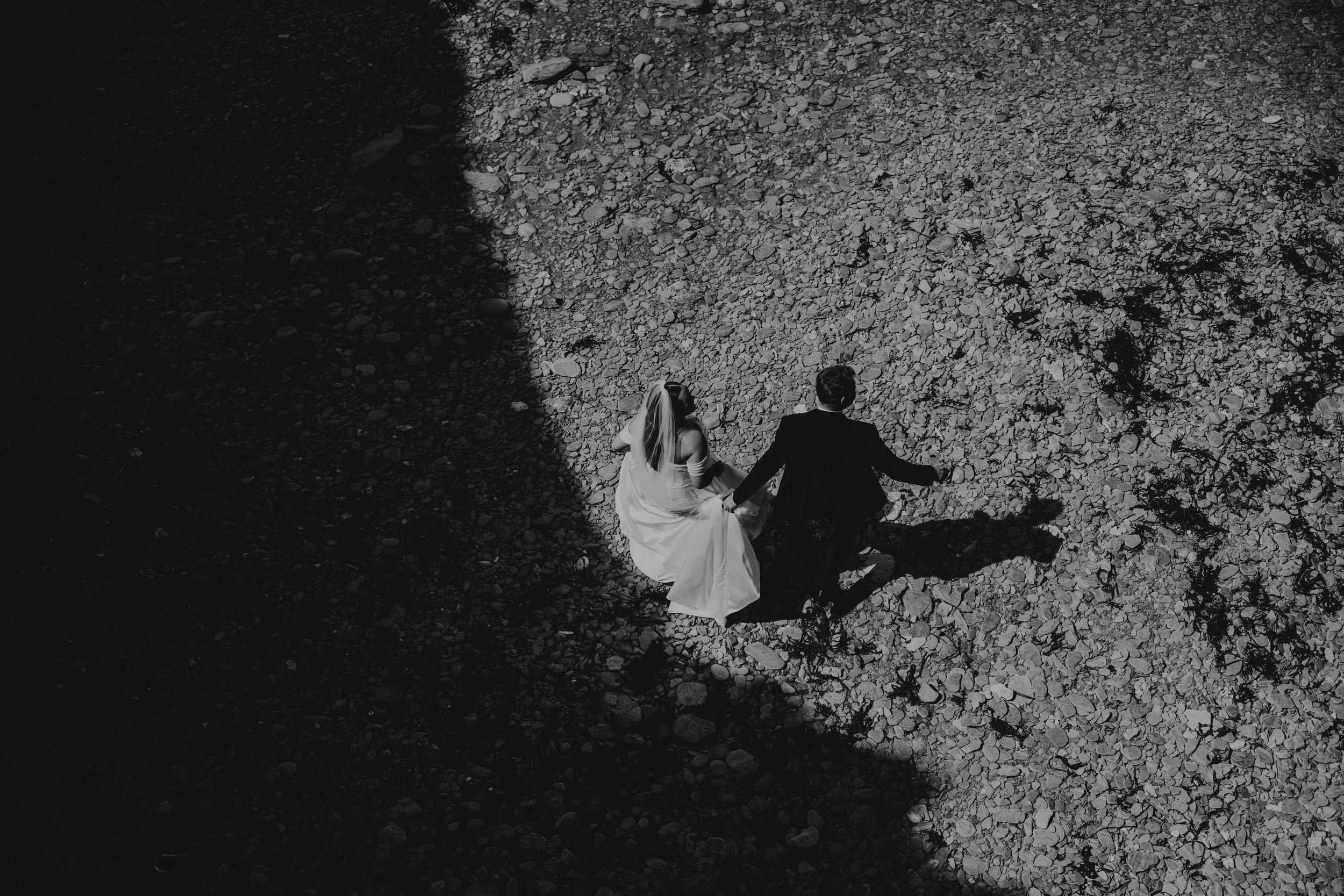 Black and white aerial view of a bride and groom walking hand in hand on a rocky beach.