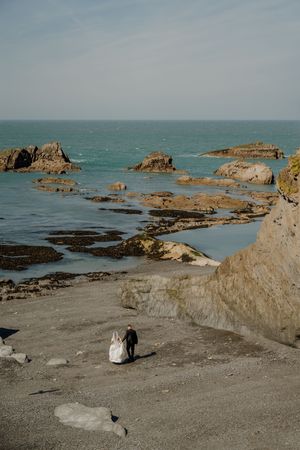 Bride and groom walk hand in hand along a dramatic rocky coastal beach after their wedding.