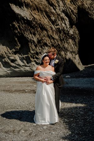 Bride and groom embrace romantically in front of dramatic rocky cliff face on their wedding day.