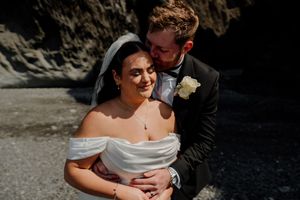 A bride and groom share a tender embrace outdoors, with the groom kissing her cheek as she smiles softly.