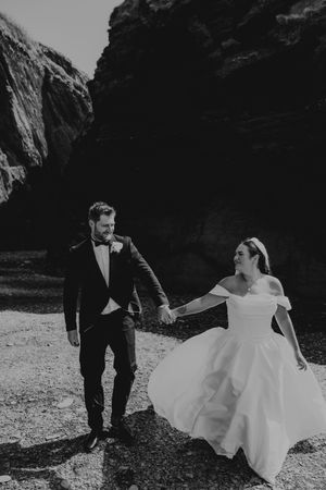 Black and white photo of bride and groom holding hands walking near dramatic rocky cliffs on their wedding day.