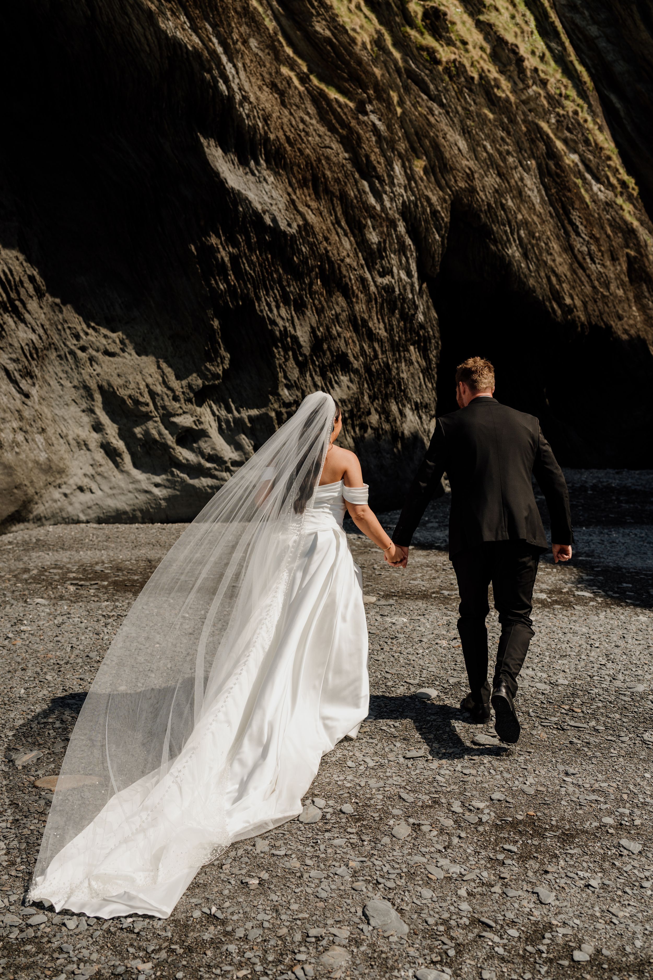 Bride in flowing white gown and veil walks hand in hand with groom toward rocky coastal cave.
