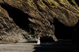 Bride in white gown poses dramatically against towering dark basalt rock columns on a black sand beach.
